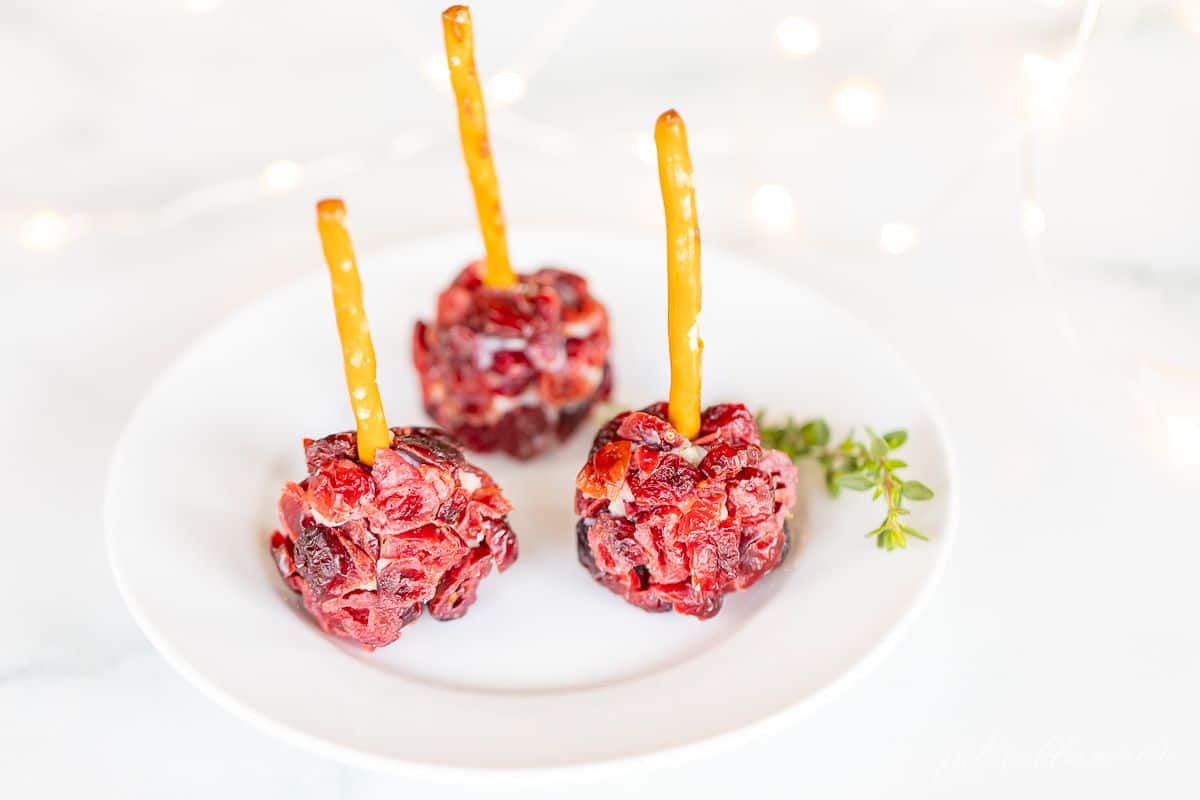 Individual cheeseball bites, covered in dried cranberries with pretzel sticks sticking out of each on a white plate.