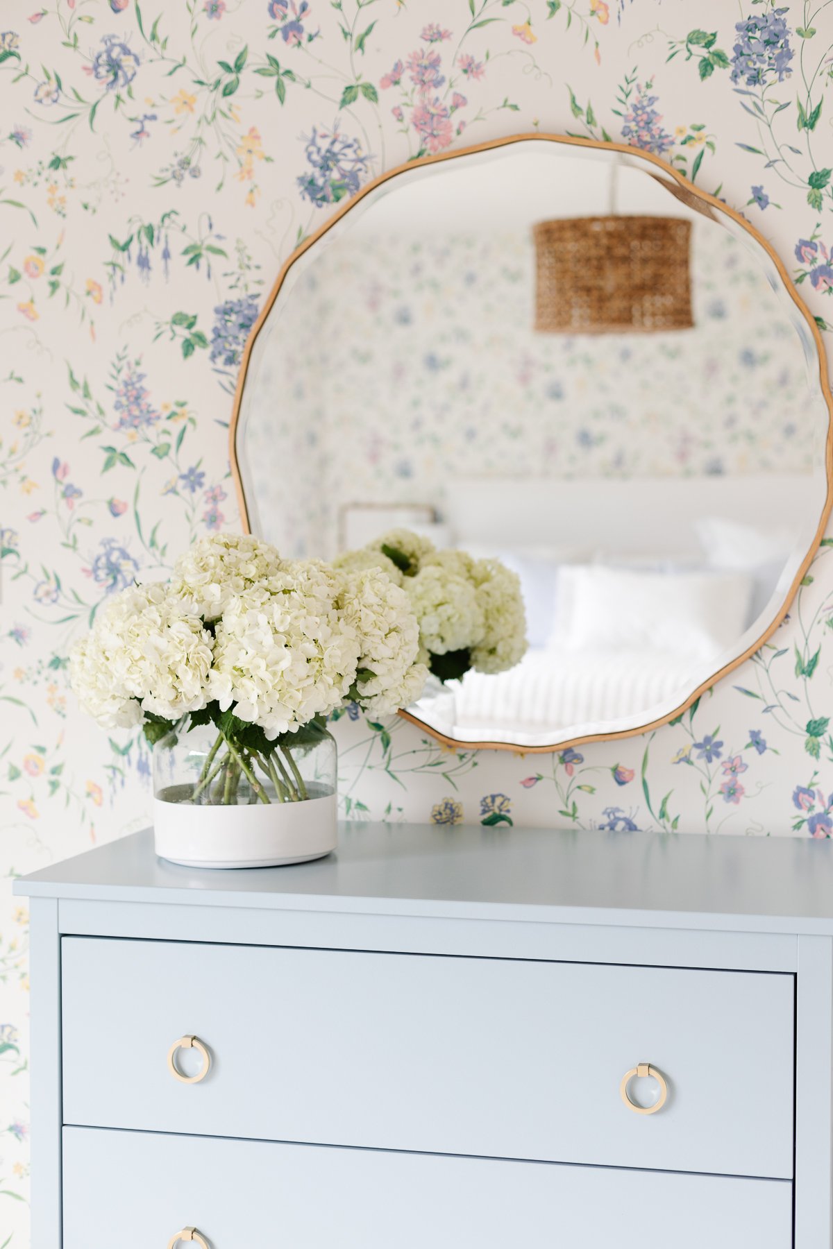 A guest bedroom with flowered wallpaper, a wavy mirror and flowers on top of a blue dresser.