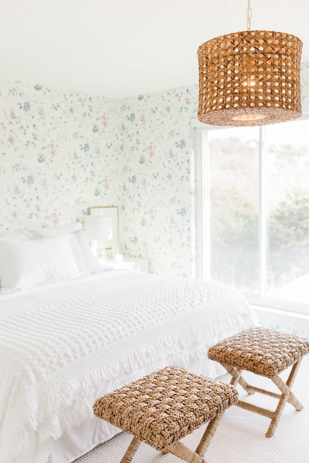 A wallpapered guest bedroom with white bedding and rattan stools.