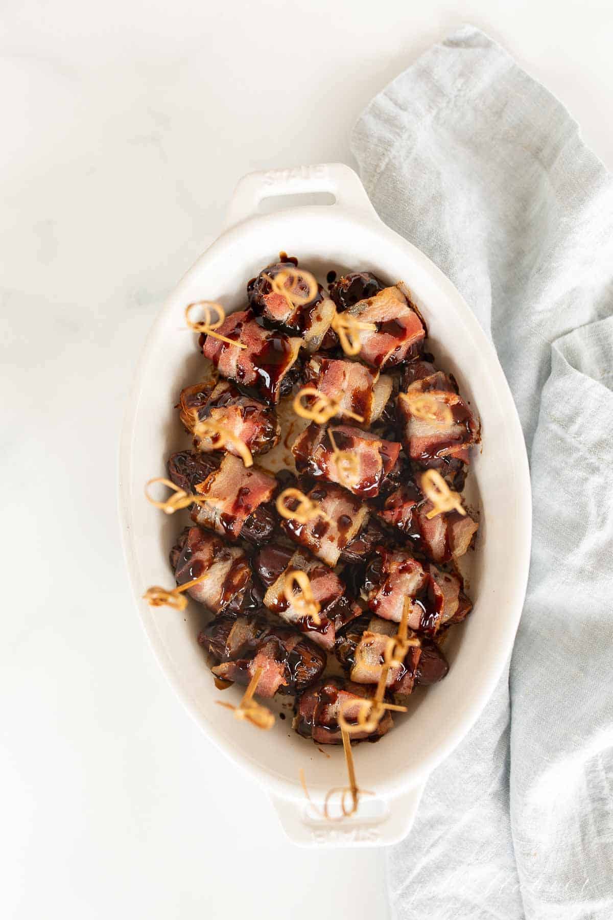 A white oval baking dish filled with dates wrapped in bacon, toothpicks in each one.