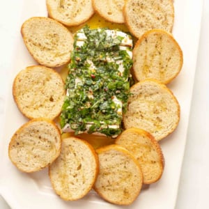 A plate of marinated cheese and bread with herbs on it.