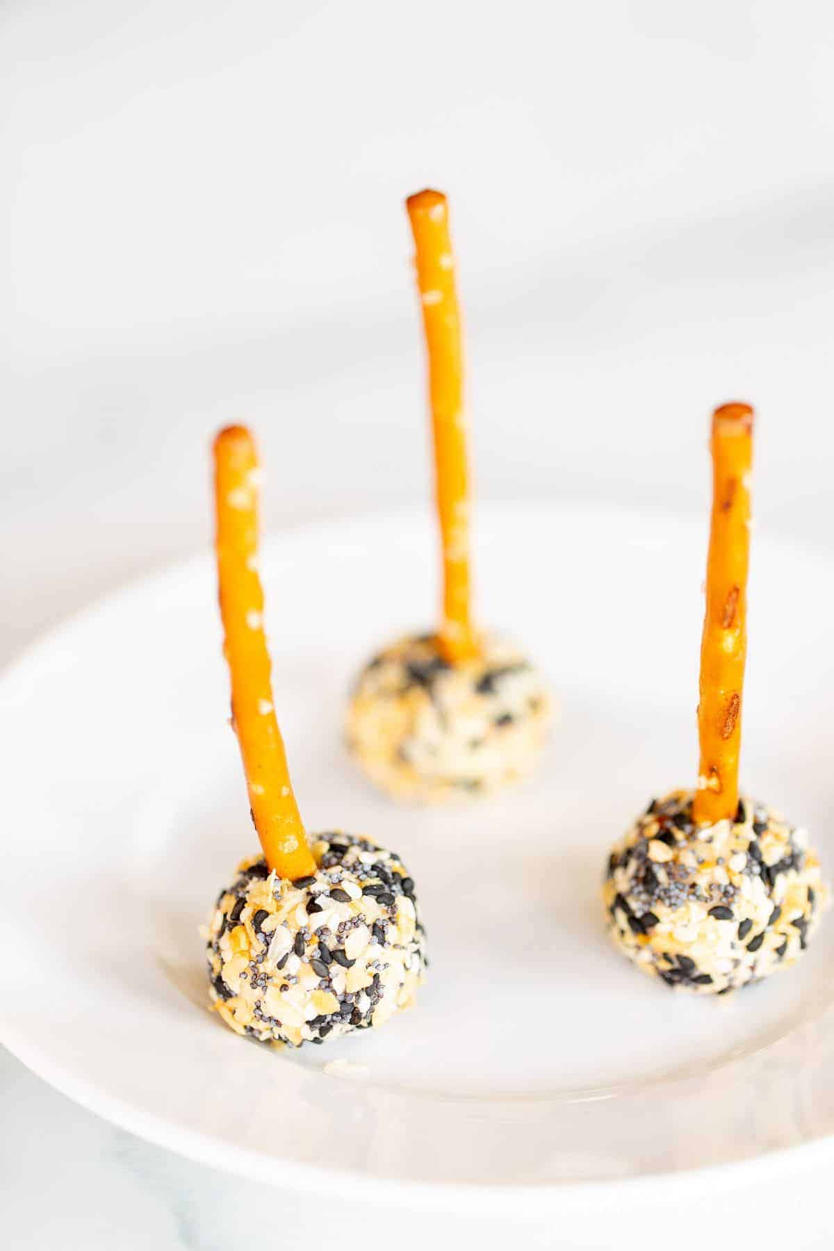 Mini cheese ball bites on a white plate, covered in everything bagel seasoning, pretzel sticks sticking out of each for serving.