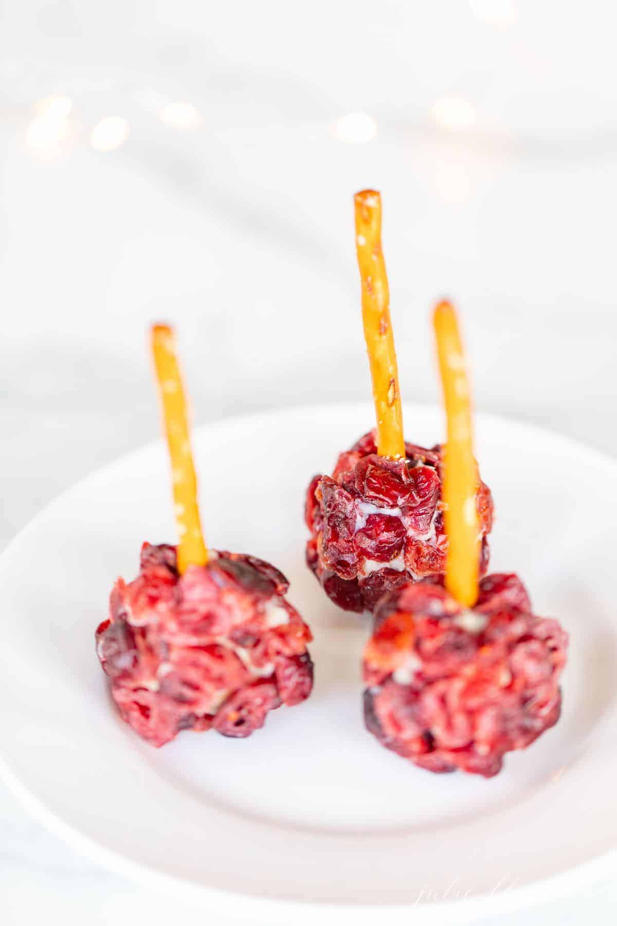 Individual cheeseball bites, covered in dried cranberries with pretzel sticks sticking out of each on a white plate.