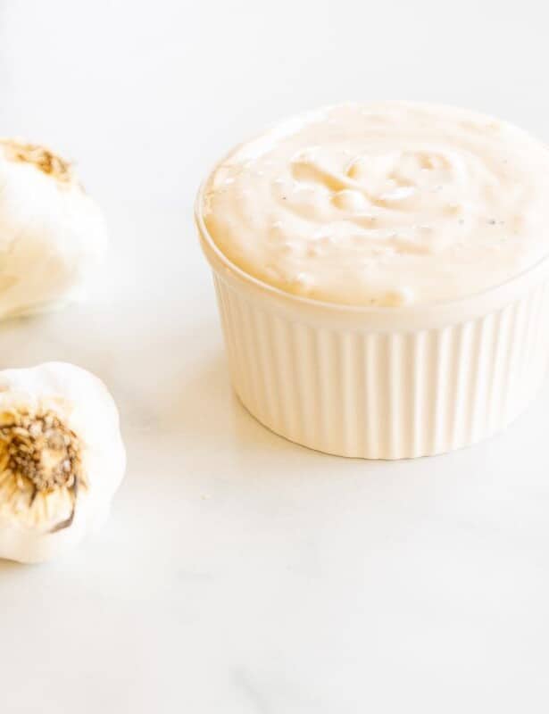 On a marble surface, a white ramekin dish full of fresh aioli, with heads of garlic to the side.