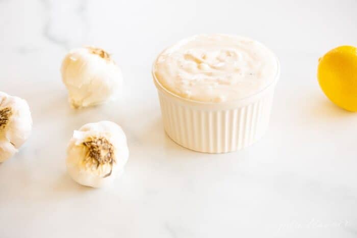On a marble surface, a white ramekin dish full of fresh aioli, with heads of garlic to the side.