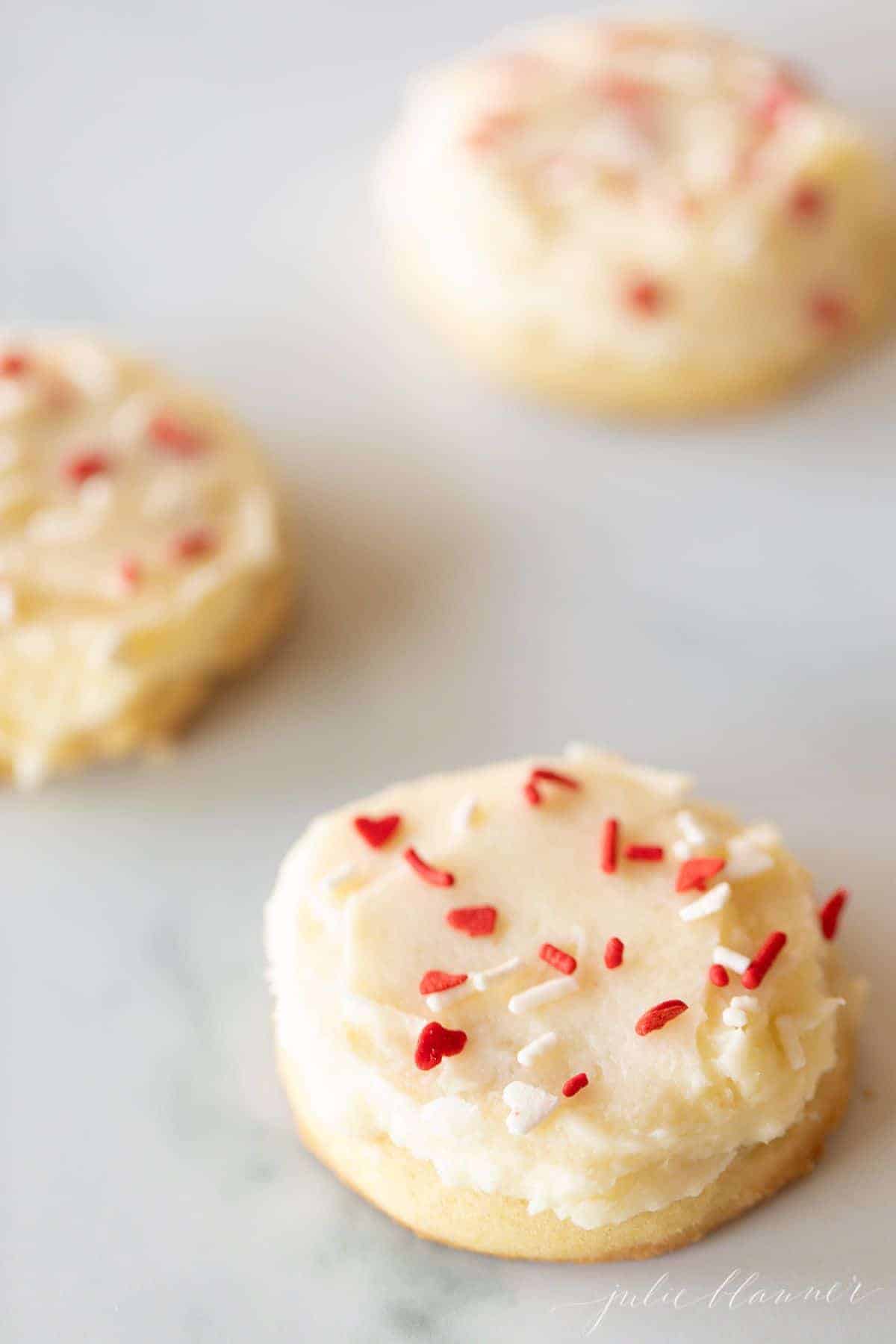 Three melt in your mouth sugar cookies frosted with Valentine's sprinkles on a marble surface.