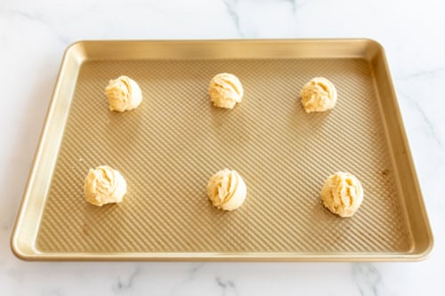 A baking sheet with six evenly spaced Amish sugar cookie dough balls rests on a marble surface.