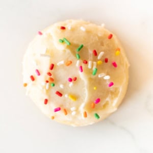 A round Amish sugar cookie topped with vanilla frosting and multi-colored sprinkles on a white background.