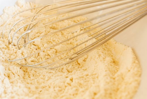 A metal whisk resting in a bowl of fine, pale yellow flour, perfect for preparing Amish sugar cookies.