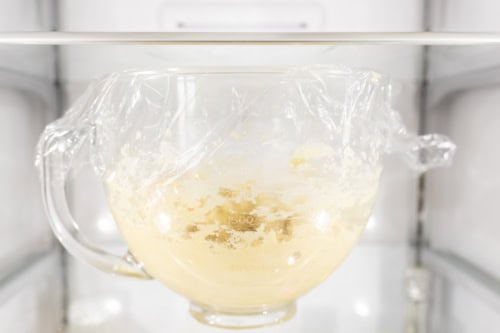 Glass bowl with dough for Amish sugar cookies covered in plastic wrap, placed on a refrigerator shelf.