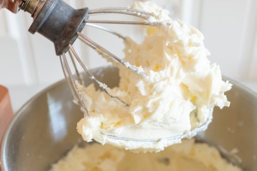 A metal whisk attachment covered with whipped buttercream frosting is held above a mixing bowl filled with more frosting, reminiscent of the creamy texture found in traditional Amish sugar cookies.