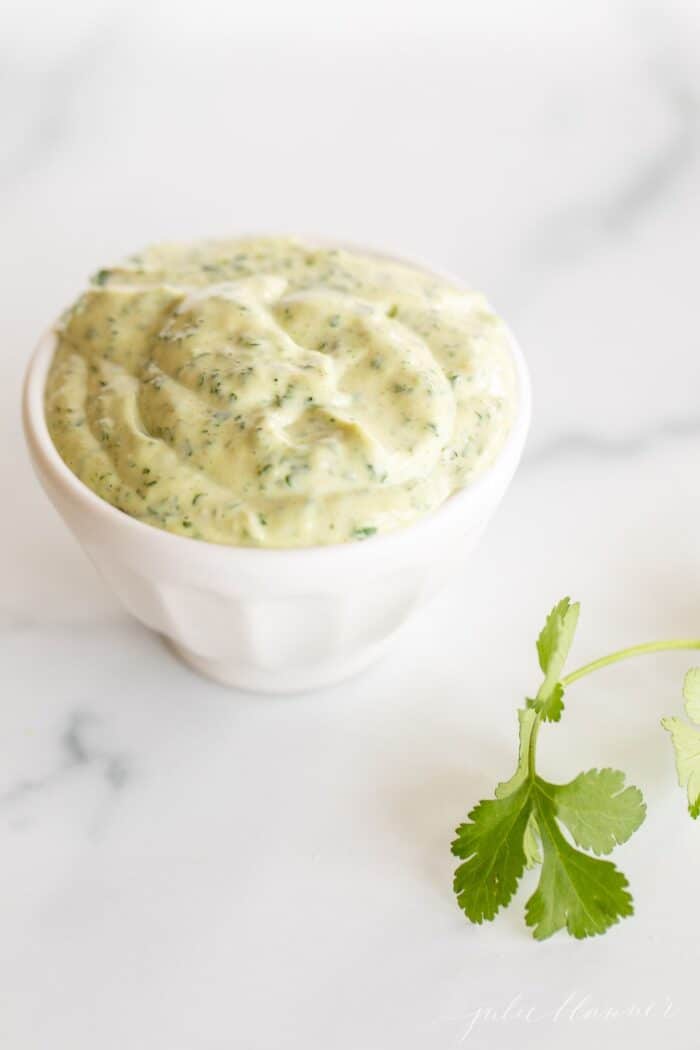 A white bowl full of cilantro lime aioli with a piece of fresh cilantro on a marble surface.