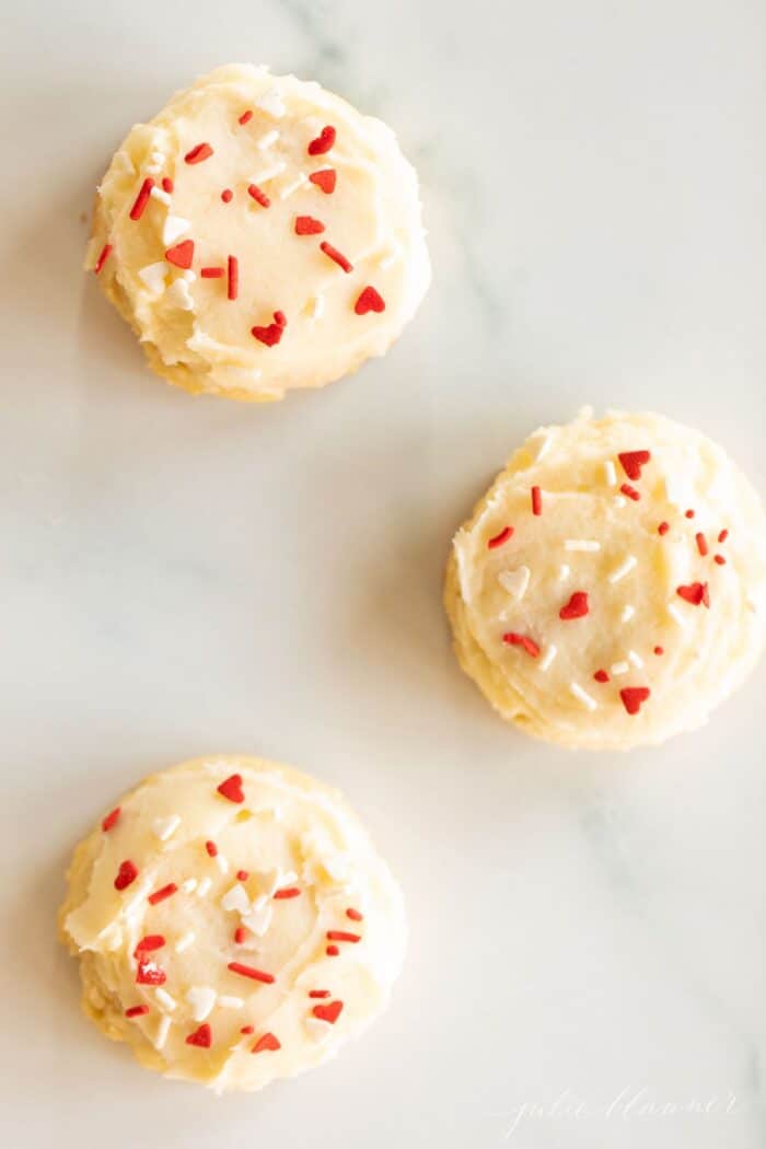 Three melt in your mouth sugar cookies frosted with Valentine's sprinkles on a marble surface.