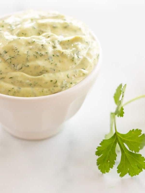 A white bowl full of cilantro lime aioli with a piece of fresh cilantro on a marble surface.