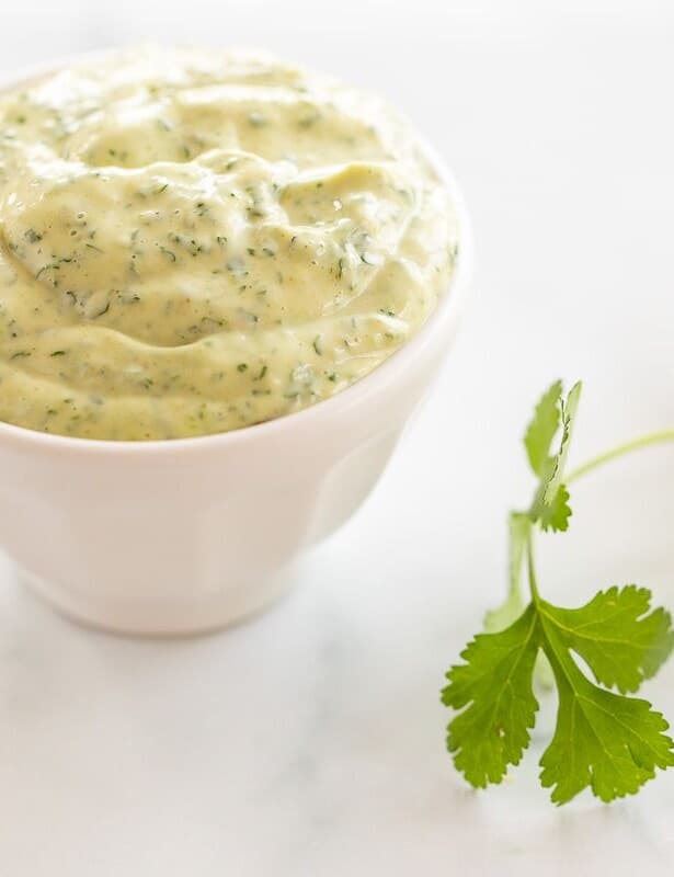 A white bowl full of cilantro lime aioli with a piece of fresh cilantro on a marble surface.