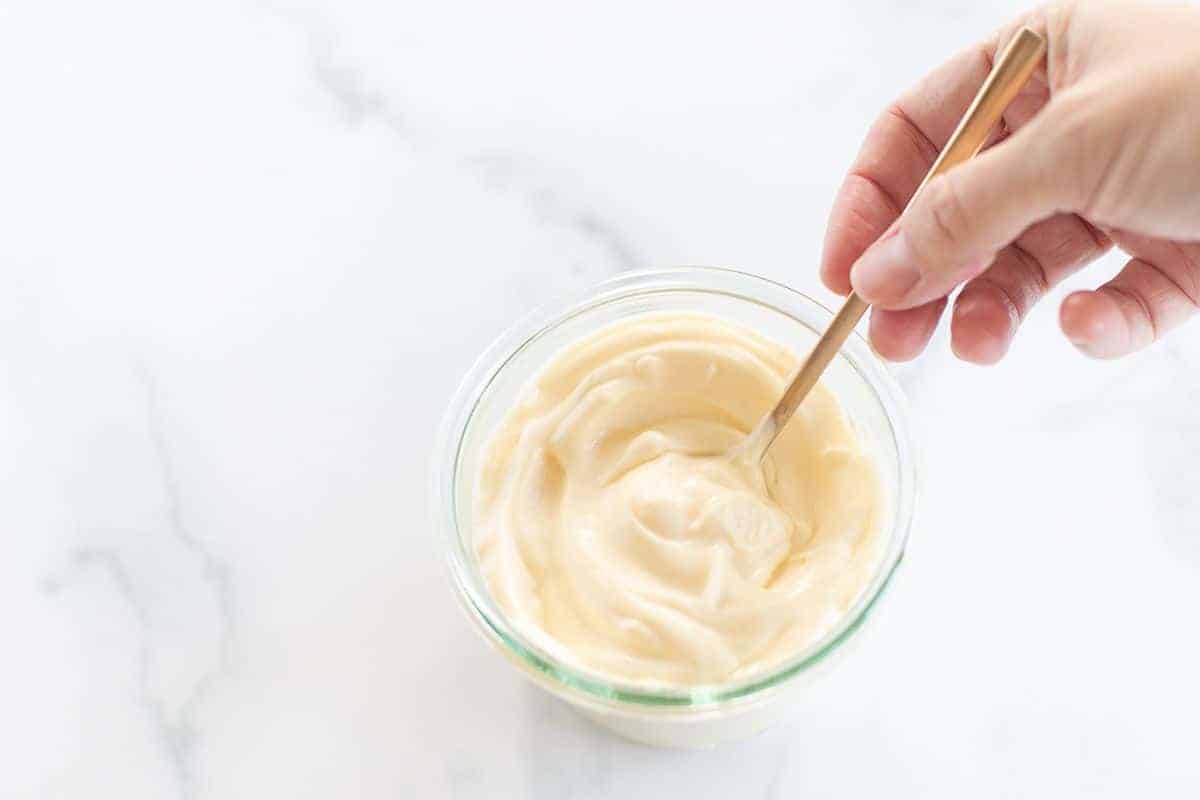 A clear glass container full of homemade mayonnaise, on a marble surface, hand holding a gold spoon inside bowl.