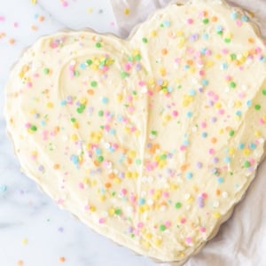 A frosted cookie cake topped with sprinkles, in a heart shaped pan, on a marble countertop.