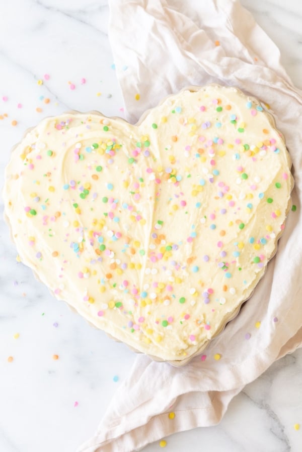 A frosted cookie cake topped with sprinkles, in a heart shaped pan, on a marble countertop.