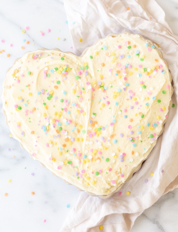 A frosted cookie cake topped with sprinkles, in a heart shaped pan, on a marble countertop.