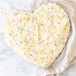 A frosted cookie cake topped with sprinkles, in a heart shaped pan, on a marble countertop.