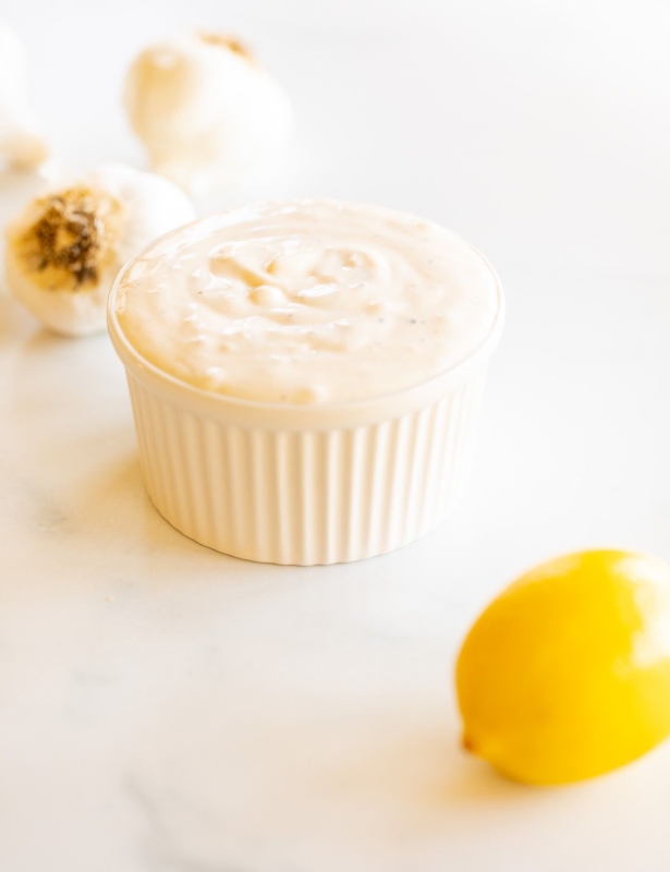 A white ramekin full of garlic aioli with a garlic bulb and a lemon in the foreground.