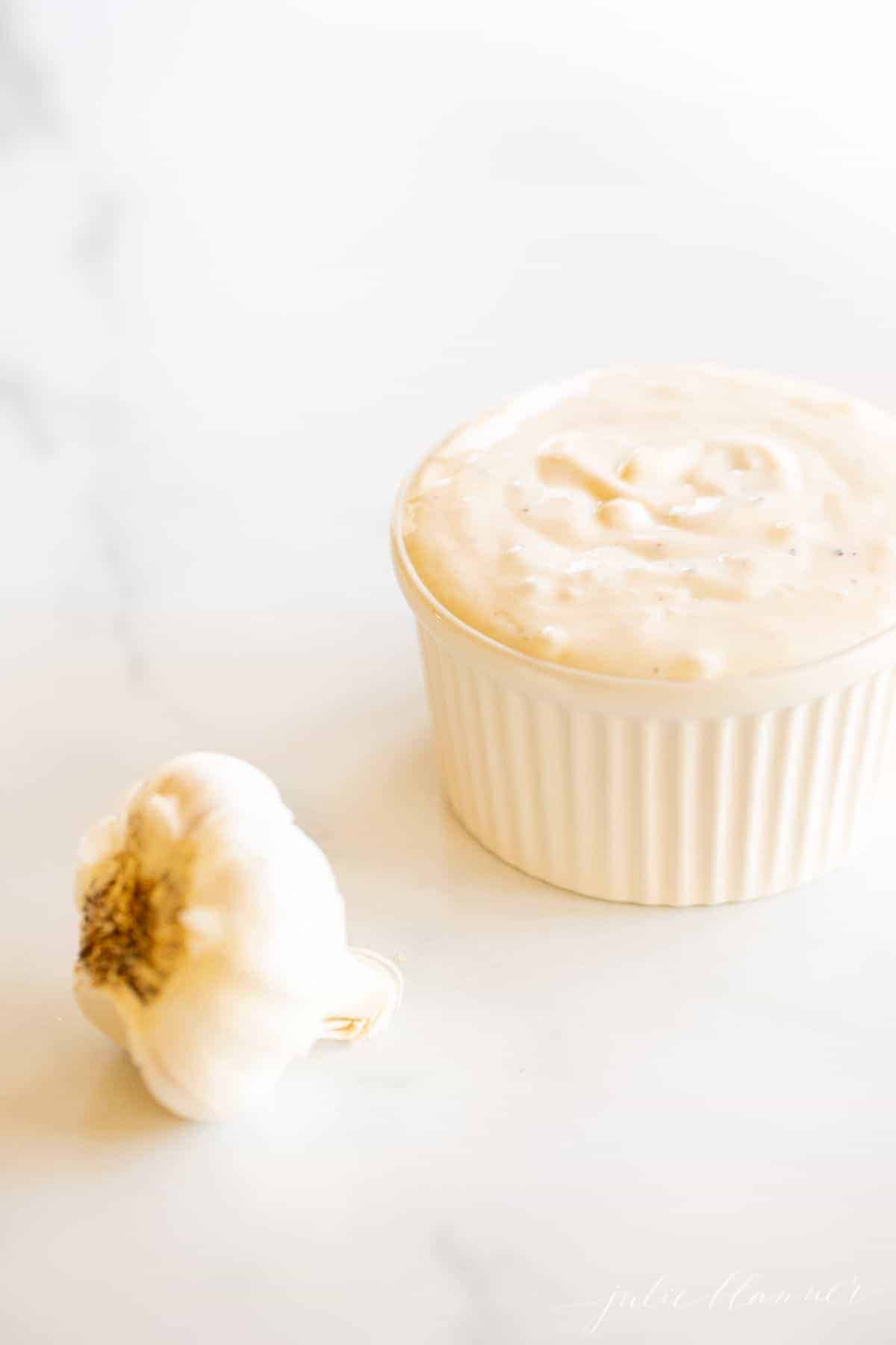 On a marble surface, a white ramekin dish full of fresh garlic aioli, with a head of garlic to the side.