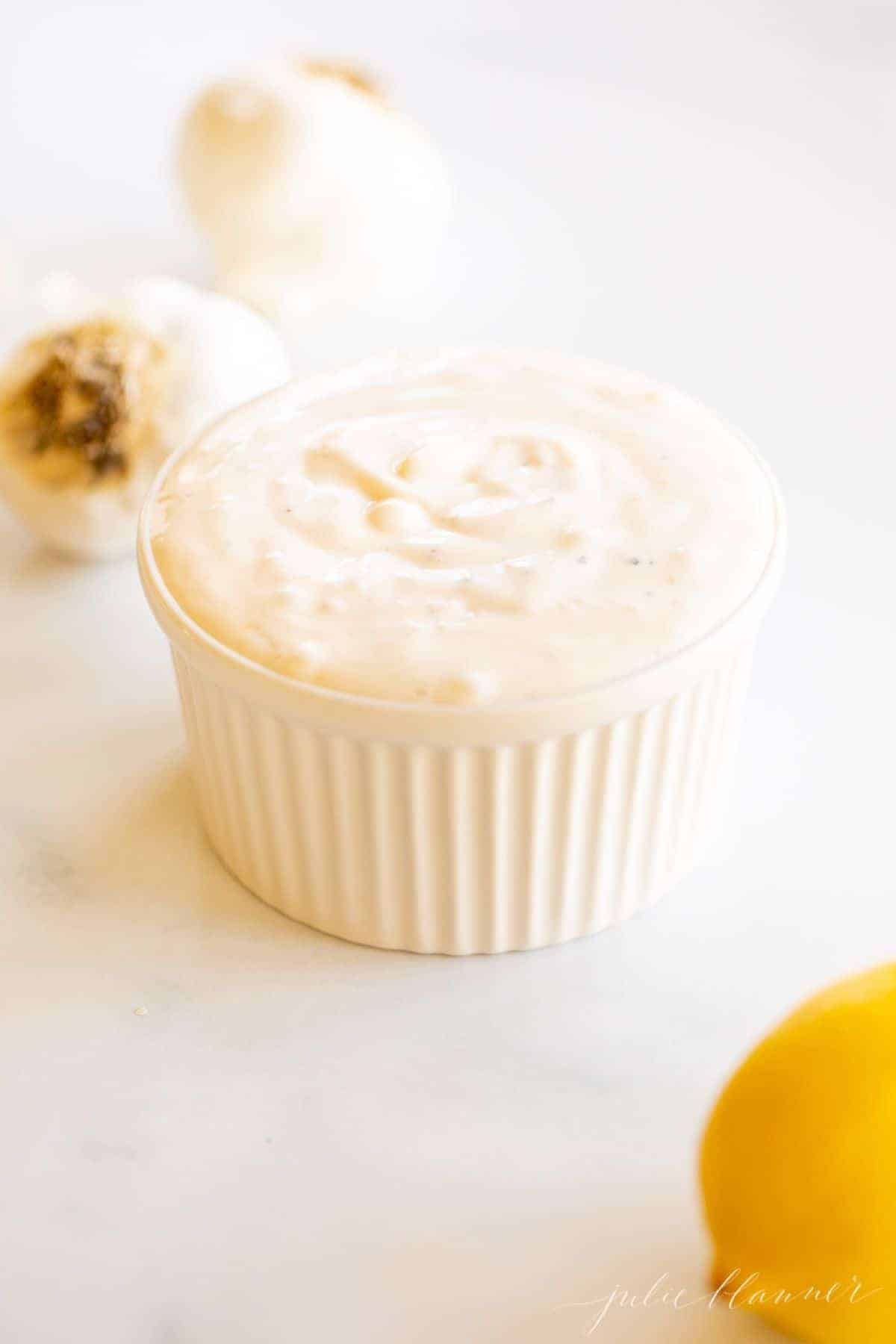 On a marble surface, a white ramekin dish full of fresh garlic aioli, with heads of garlic and a lemon to the side.