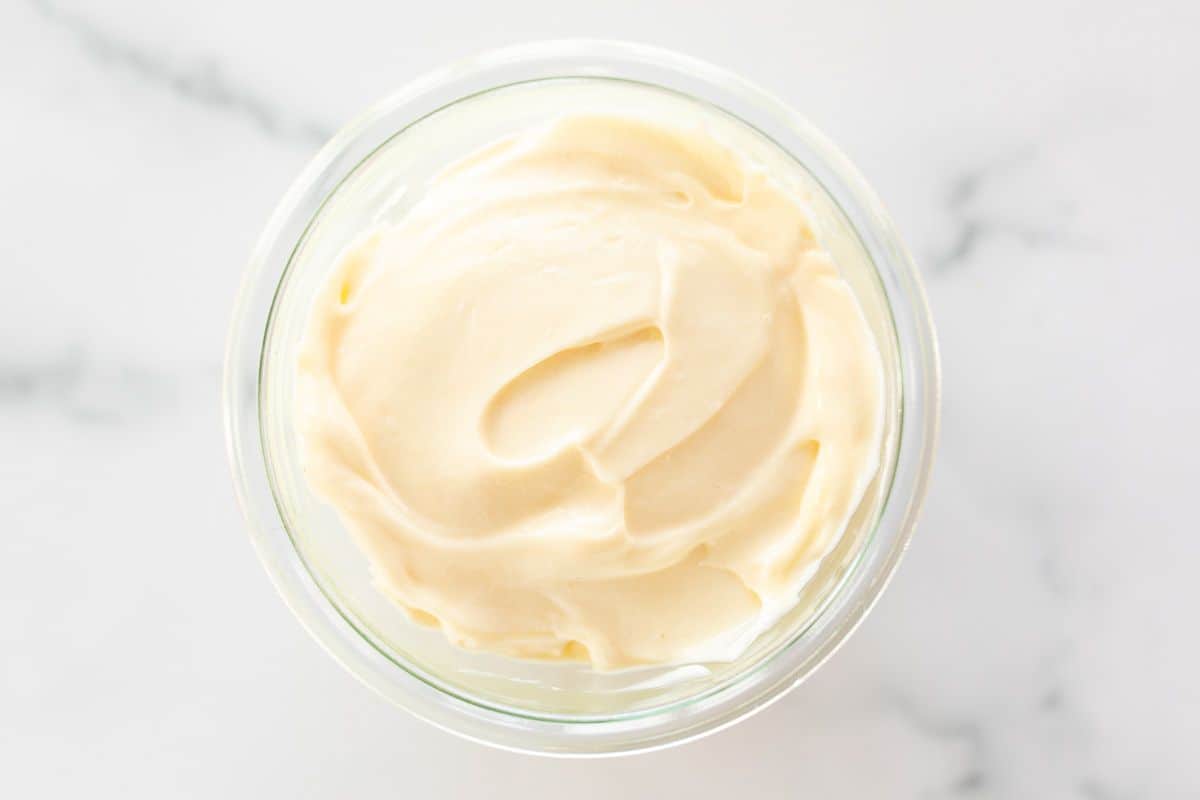A clear glass container full of homemade mayonnaise, on a marble surface.