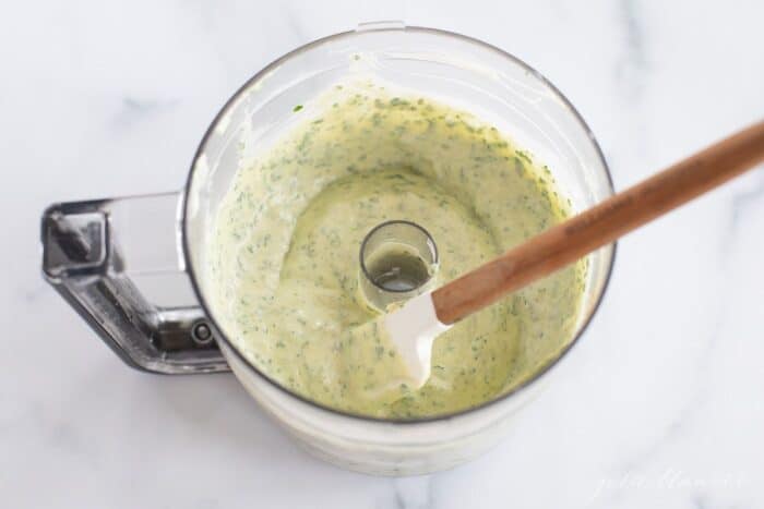The bowl of a food processor filled with fresh cilantro aioli, wooden spatula to the side.
