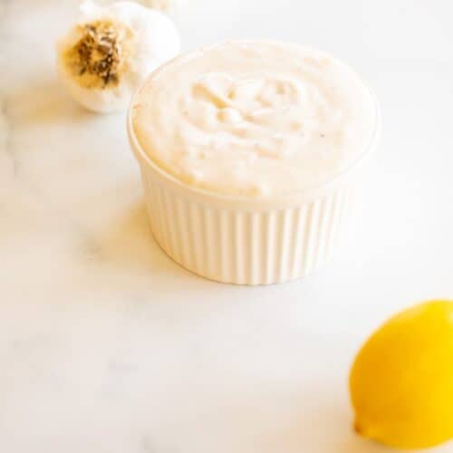 On a marble surface, a white ramekin dish full of fresh garlic aioli, with heads of garlic and a lemon to the side.
