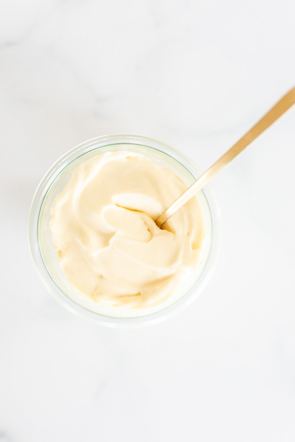 A small glass container of homemade mayonnaise with a gold spoon, resting on a marble countertop.