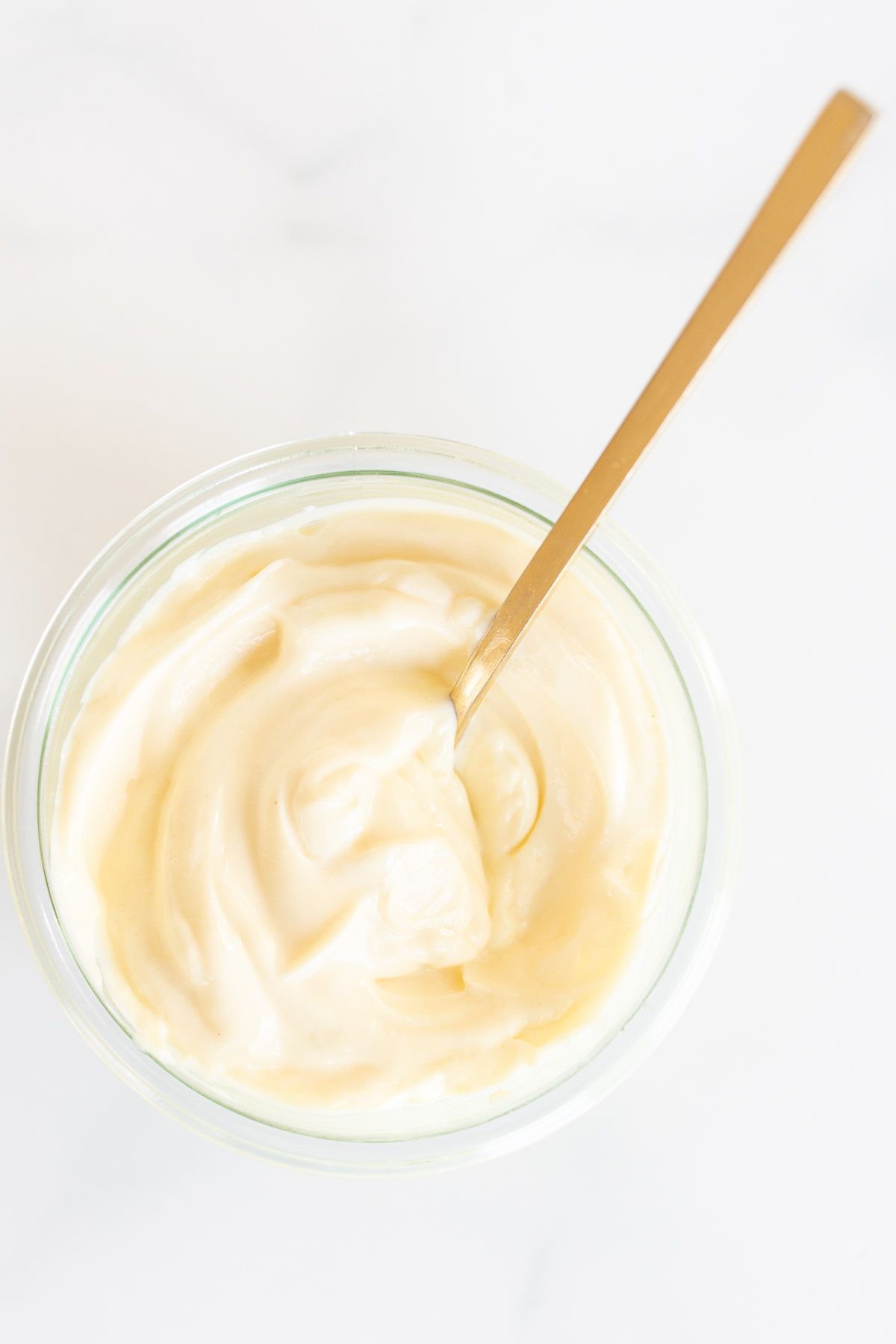 A small glass container of homemade mayonnaise with a gold spoon, resting on a marble countertop.