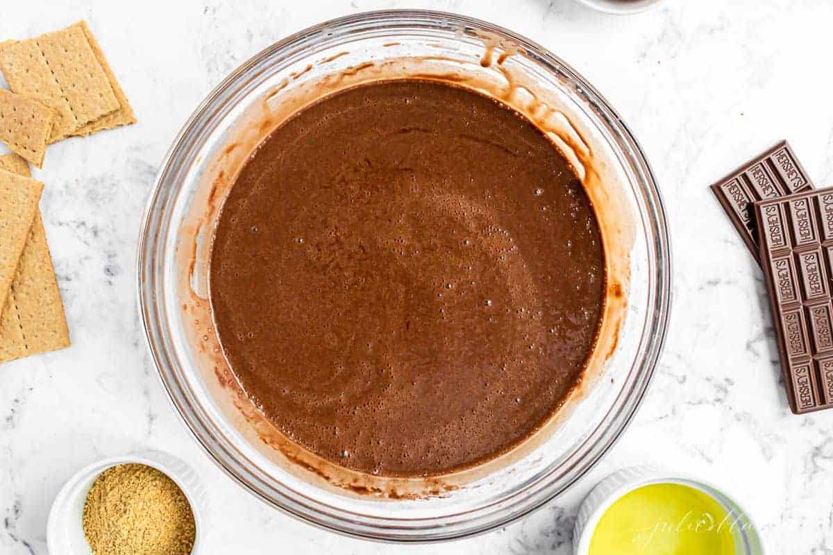Chocolate cupcake batter in a clear glass bowl, ingredients for smores cupcakes surrounding.
