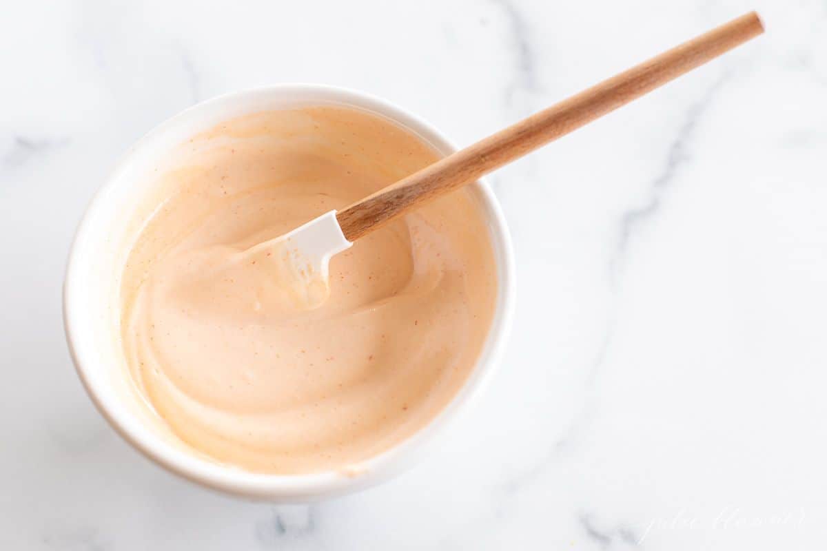 A white bowl full of homemamde sriracha mayo on a white marble background, wooden spatula in the bowl.
