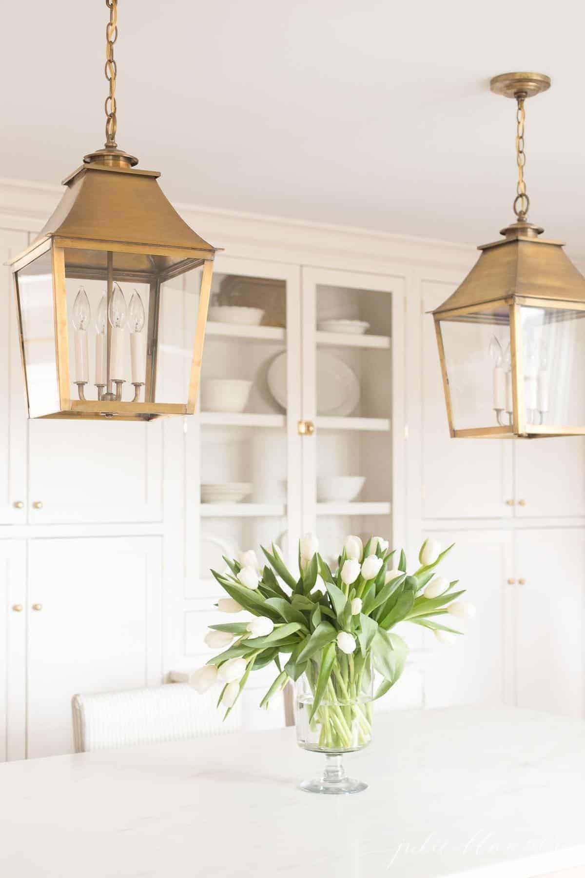 Inset cabinets in a cream kitchen with brass lanterns and a vase of flowers on the island.