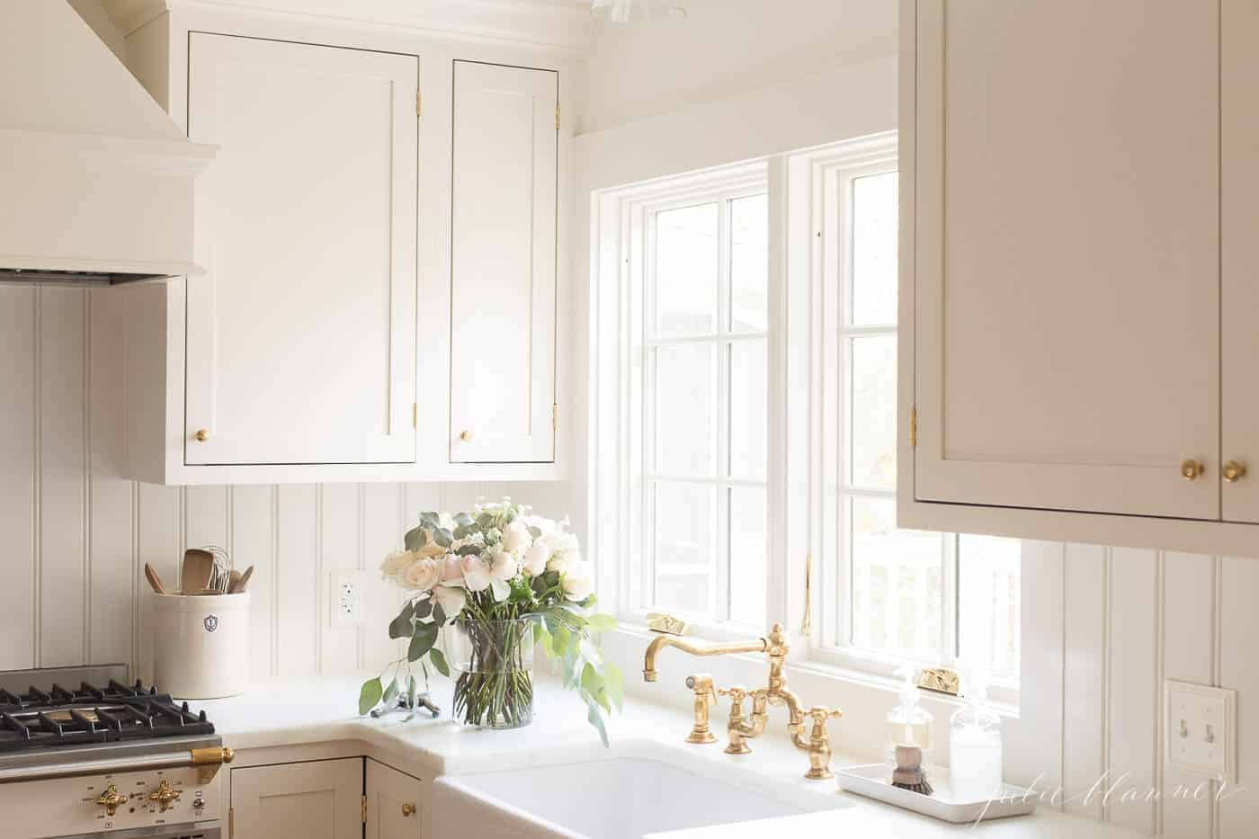 Cream inset cabinets in a kitchen with marble counters and a brass faucet.