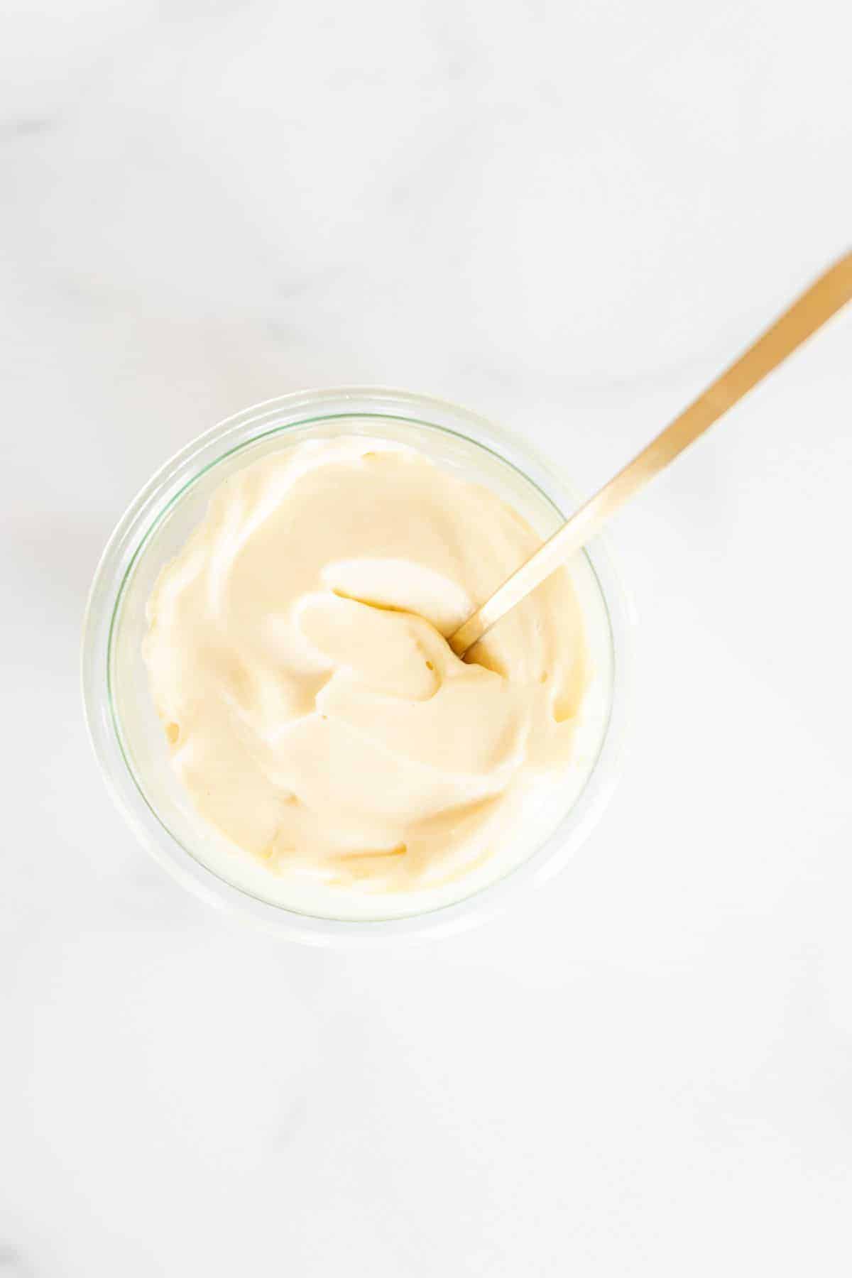 A clear glass container full of homemade mayonnaise, on a marble surface, gold spoon inside bowl.