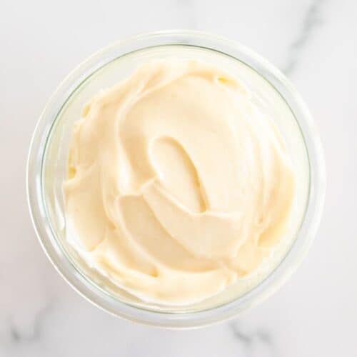 A clear glass container full of homemade mayonnaise, on a marble surface.