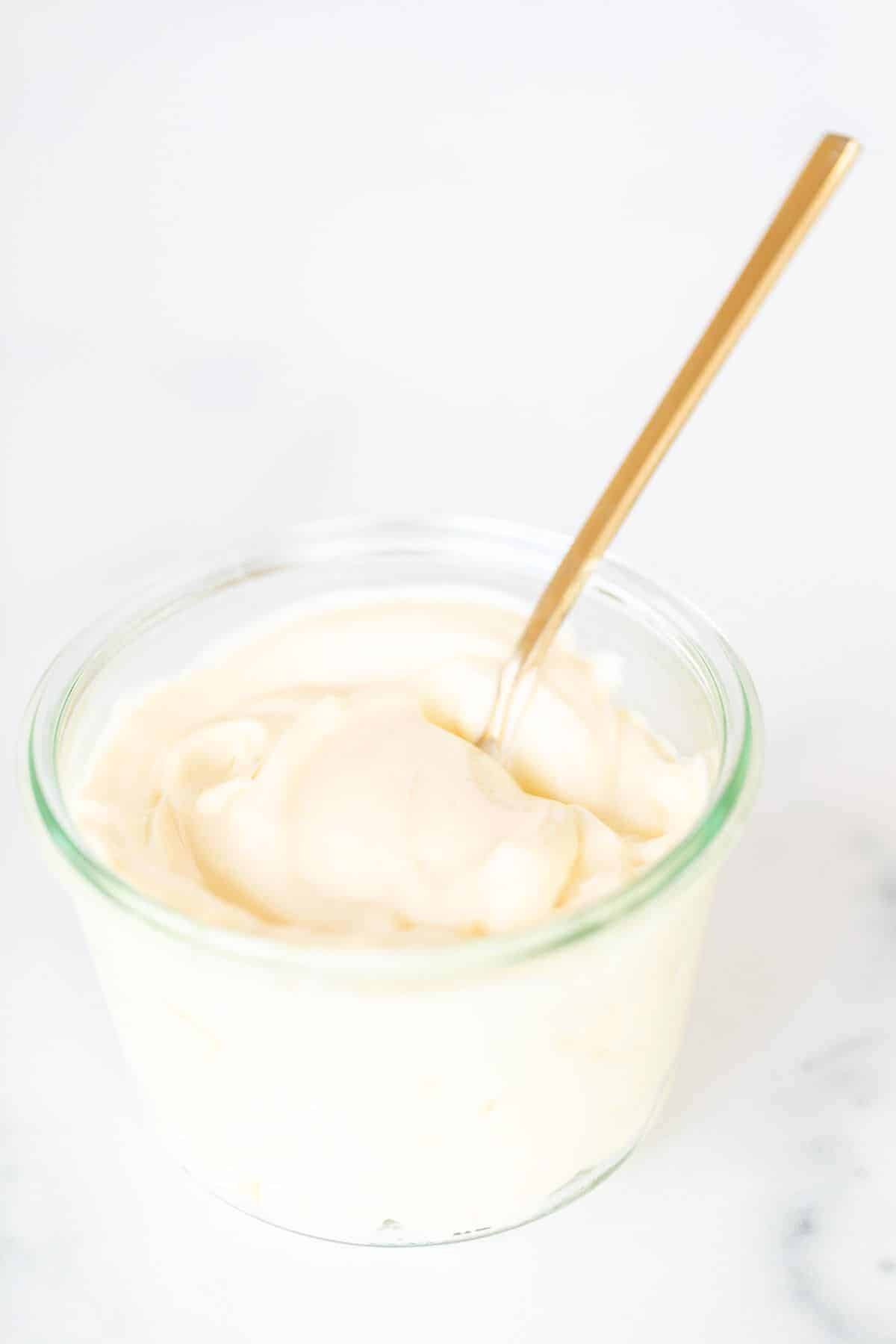 A clear glass container full of homemade mayonnaise, on a marble surface, gold spoon inside bowl.