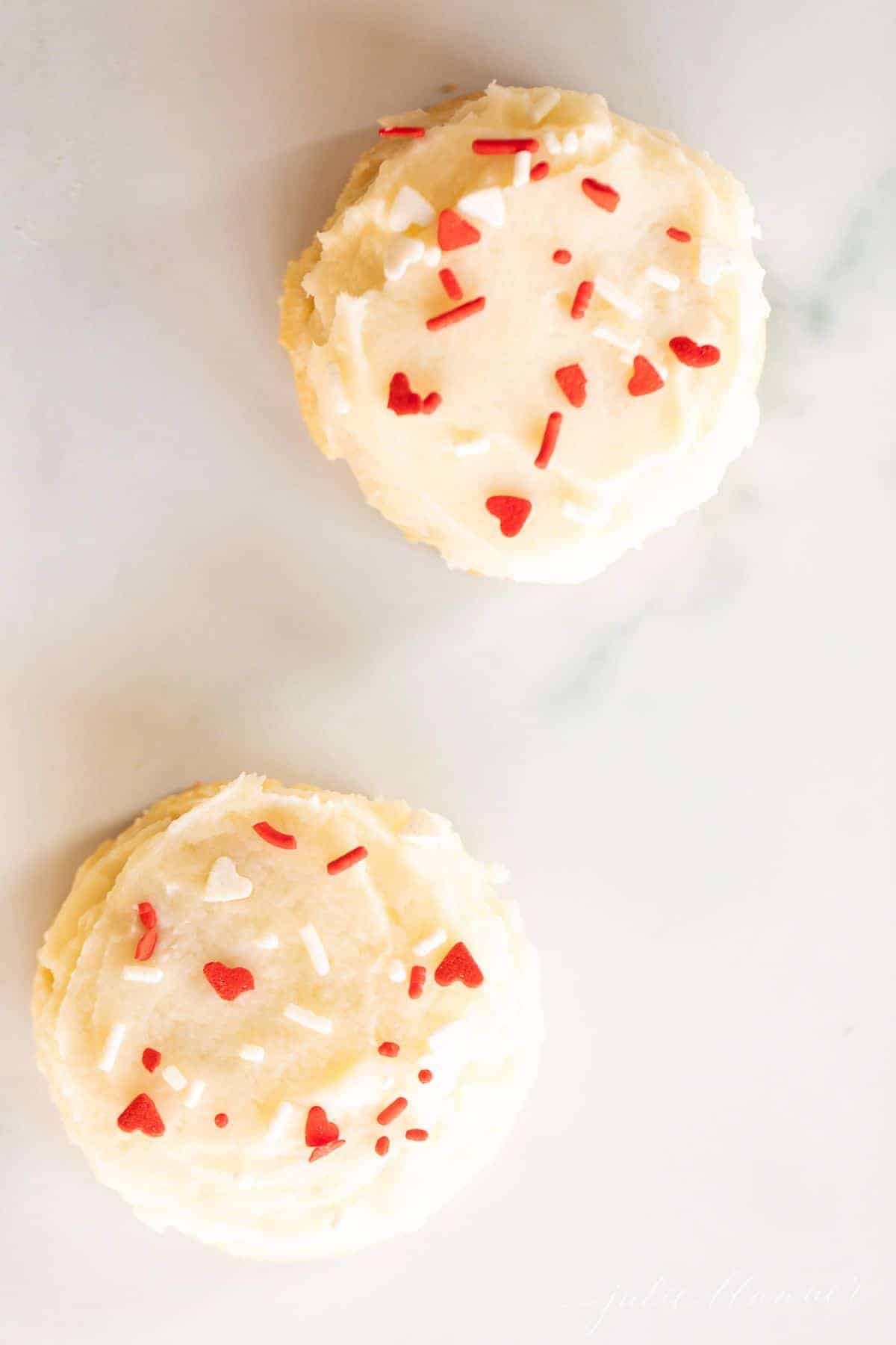 Two melt in your mouth sugar cookies frosted with Valentine's sprinkles on a marble surface.