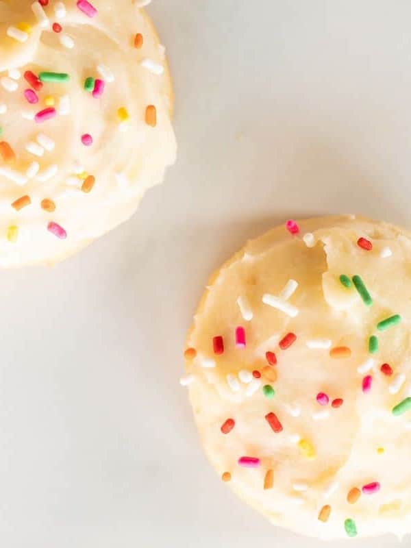 Two melt in your mouth sugar cookies frosted with sprinkles on a marble surface.