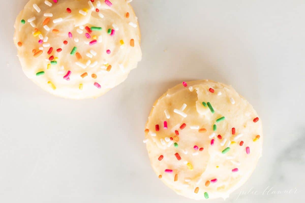 Two melt in your mouth sugar cookies frosted with sprinkles on a marble surface.