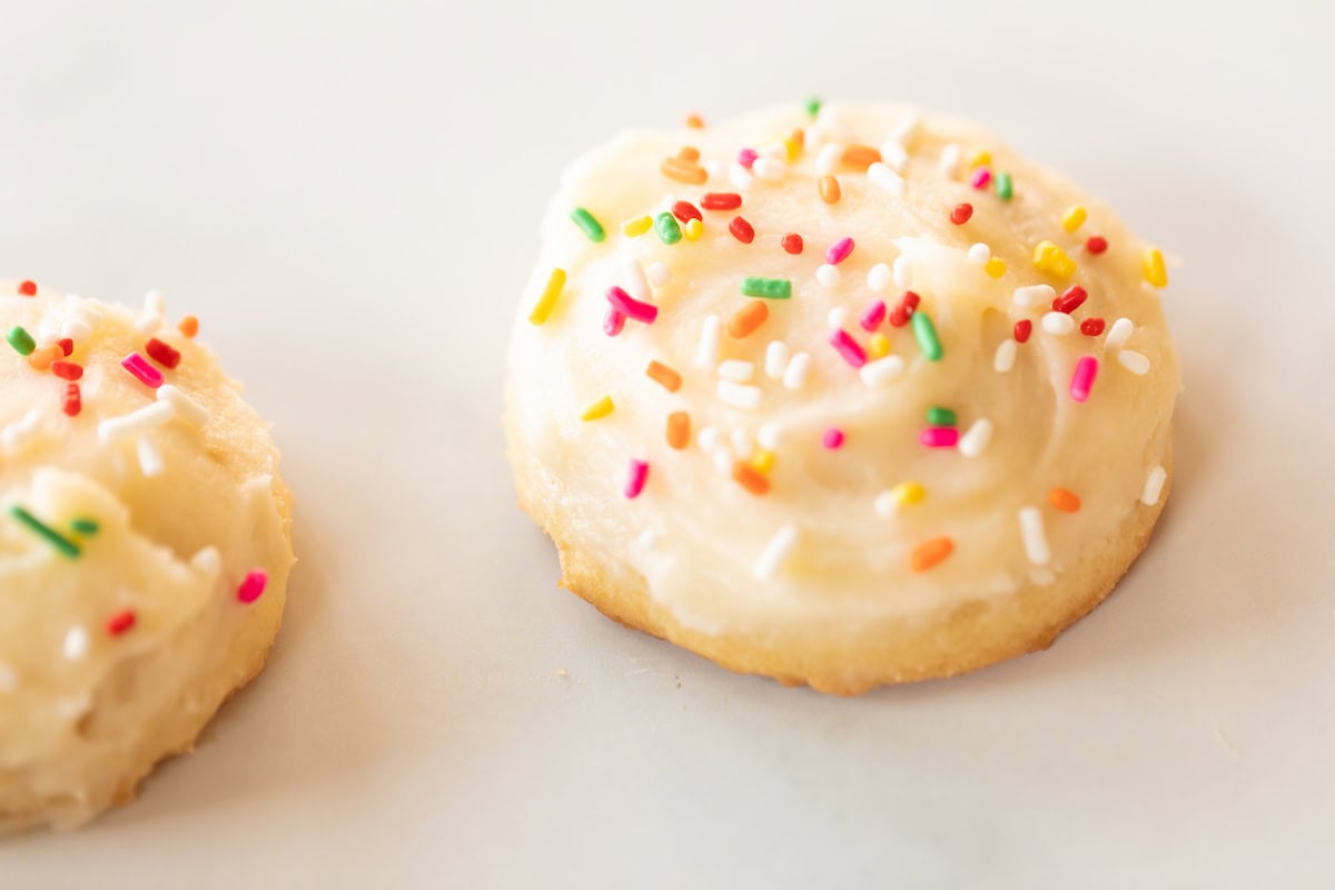 A frosted melt in your mouth sugar cookie with colorful sprinkles sits elegantly on a plain white surface.