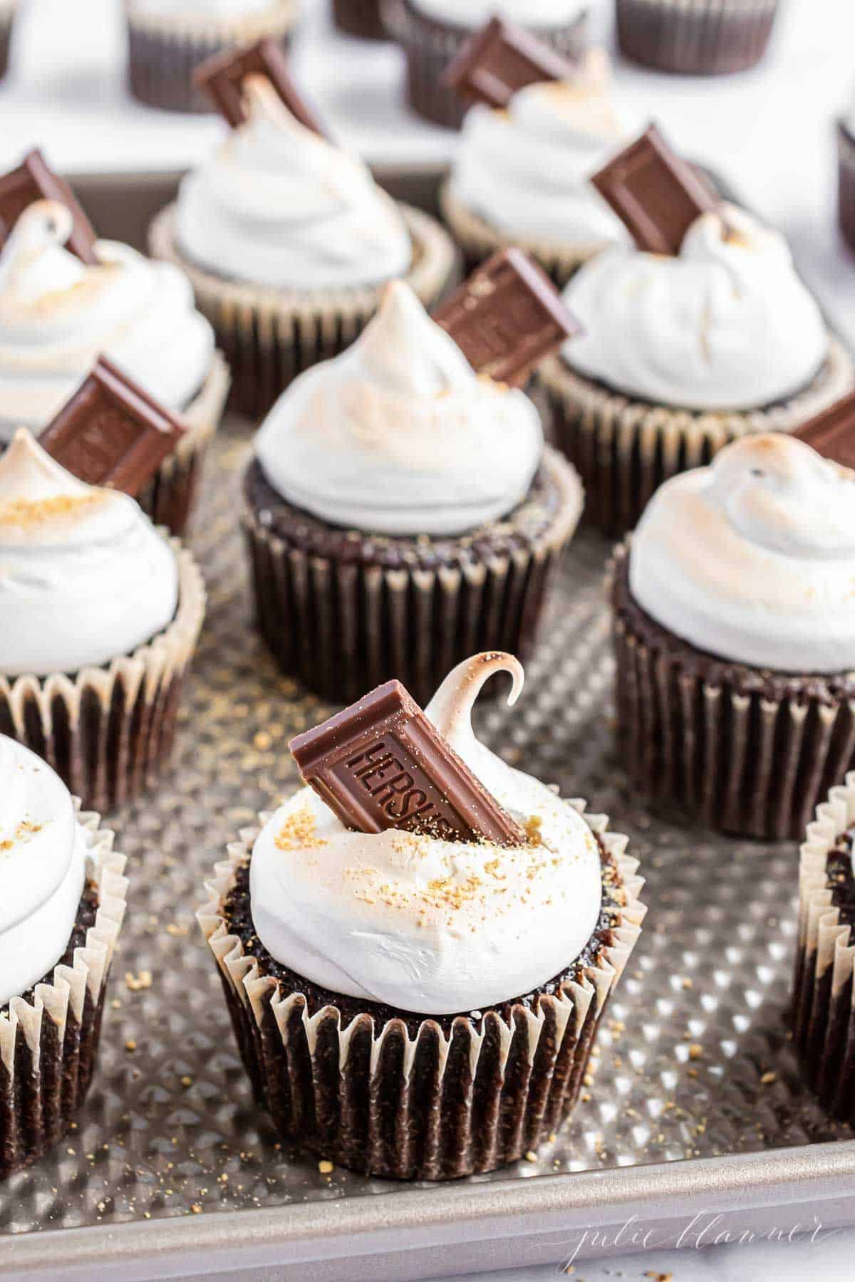Homemade smores cupcakes on a baking sheet, lined up and topped with marshmallow frosting and a piece of Hersheys bar.