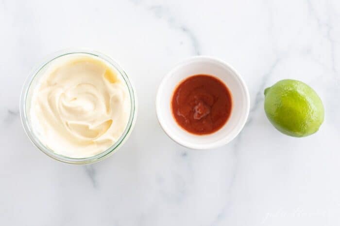 A bowl of mayonnaise, a bowl of sriracha and a whole lime, sitting in a row on a marble surface.