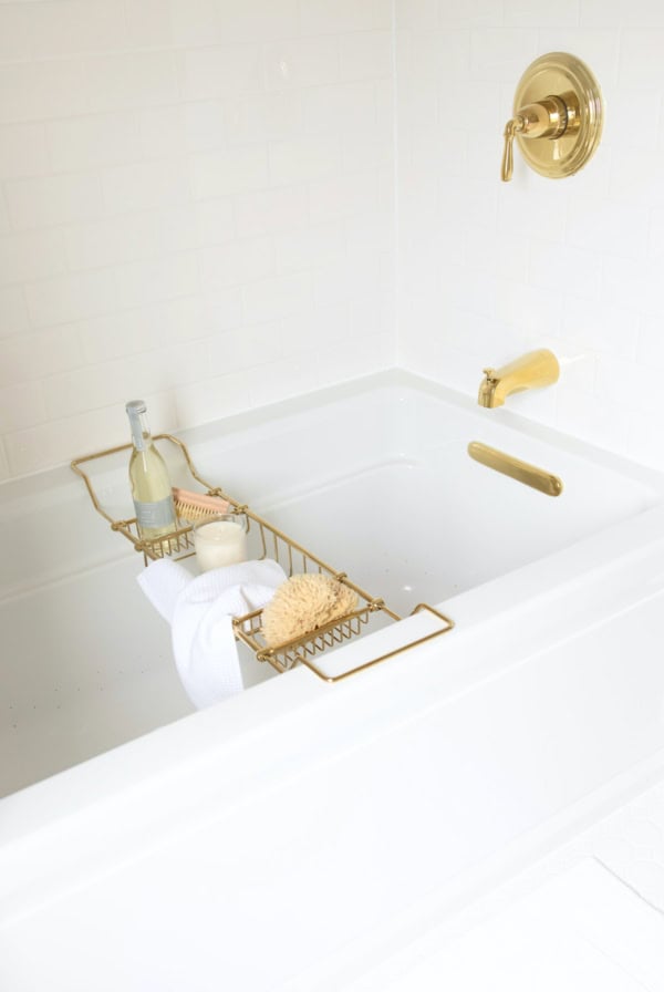 A white bathtub with gold fixtures sits against a backdrop of subway tile. A gold bath caddy holds a bottle of wine, a bath sponge, and a folded white towel.