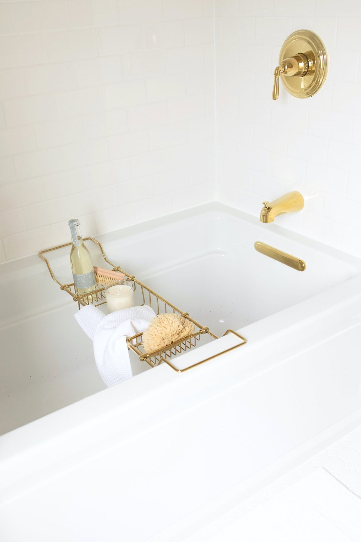 A white bathtub with gold fixtures sits against a backdrop of subway tile. A gold bath caddy holds a bottle of wine, a bath sponge, and a folded white towel.