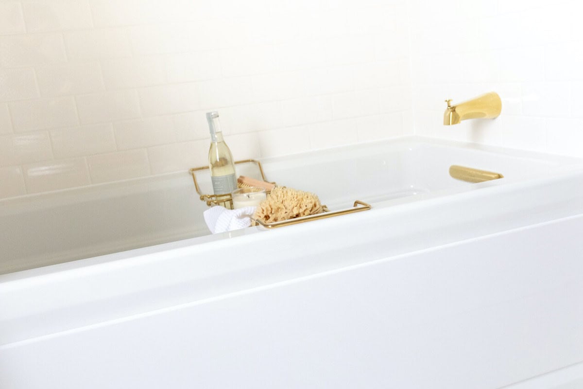 A white bathtub with a gold faucet sits beneath gleaming subway tiles, accompanied by a bath caddy holding a bottle, a sponge, and a towel.