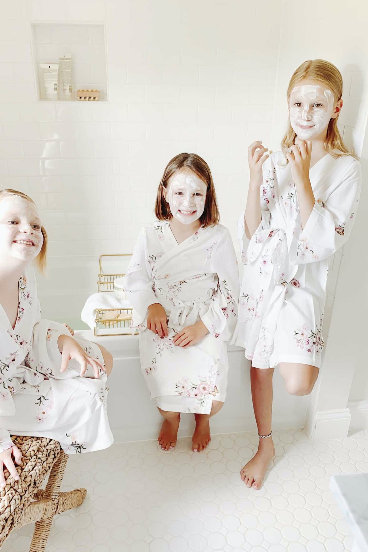 Three children in floral bathrobes with white face masks stand and sit in a bright bathroom adorned with subway tile. One child stands near the tub, one sits on the edge, and another sits on a wicker stool.