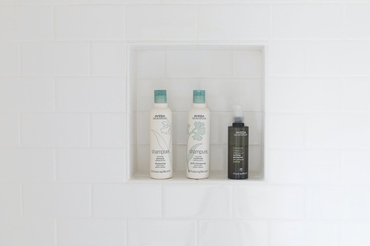 Three bathroom products, including two bottles of shampoo and one bottle of conditioner, are neatly arranged in a recessed shower shelf against a white subway tiled wall.